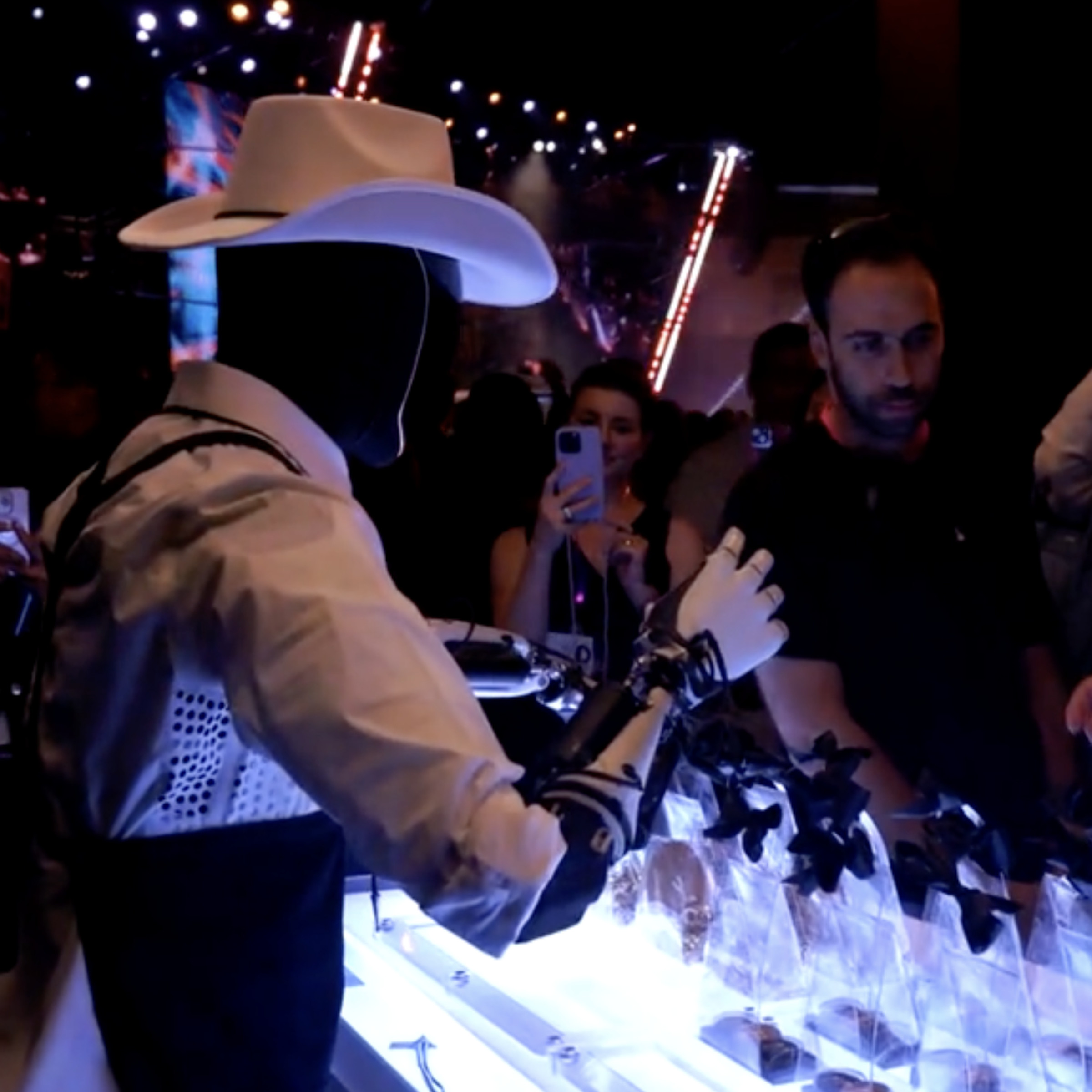 A picture of an Optimus robot, wearing a cowboy hat and standing in front of a crowd of people, behind a table.