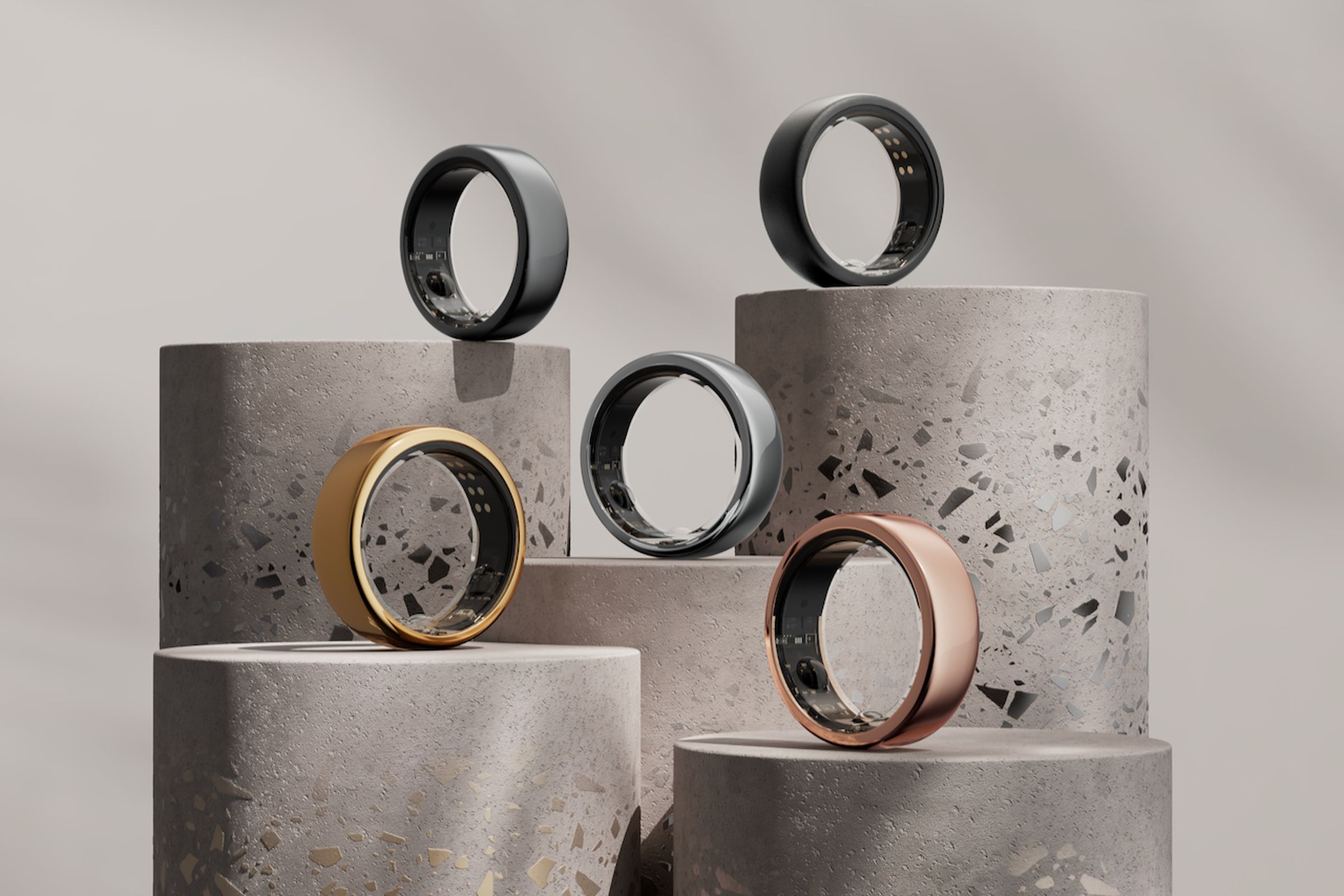 A collection of five Oura smart rings are arranged on concrete pedestals of varying heights. There’s a glossy gold ring, a rosy gold ring, a silver one in the center, a gunmetal gray one, and a matte black one.