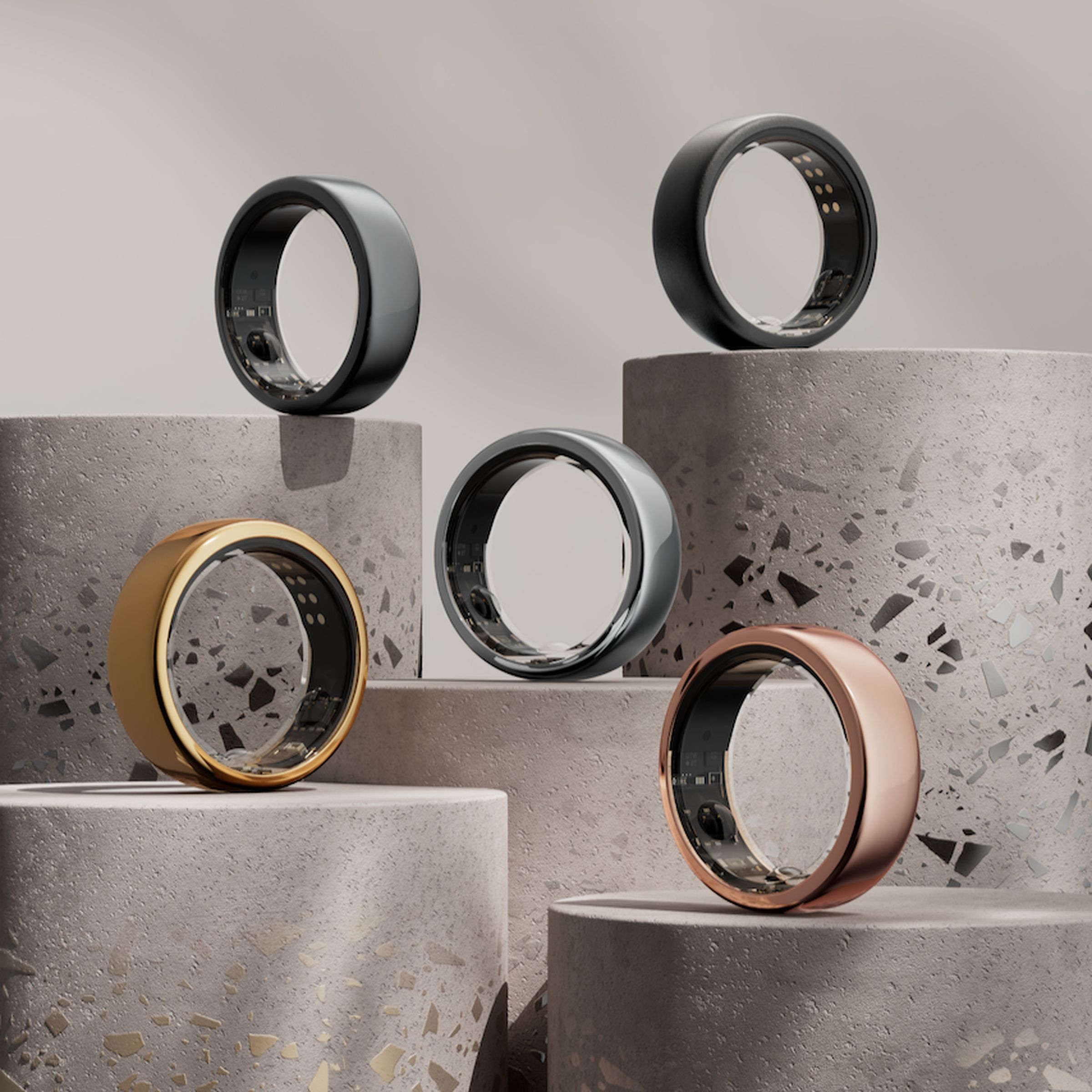 A collection of five Oura smart rings are arranged on concrete pedestals of varying heights. There’s a glossy gold ring, a rosy gold ring, a silver one in the center, a gunmetal gray one, and a matte black one.