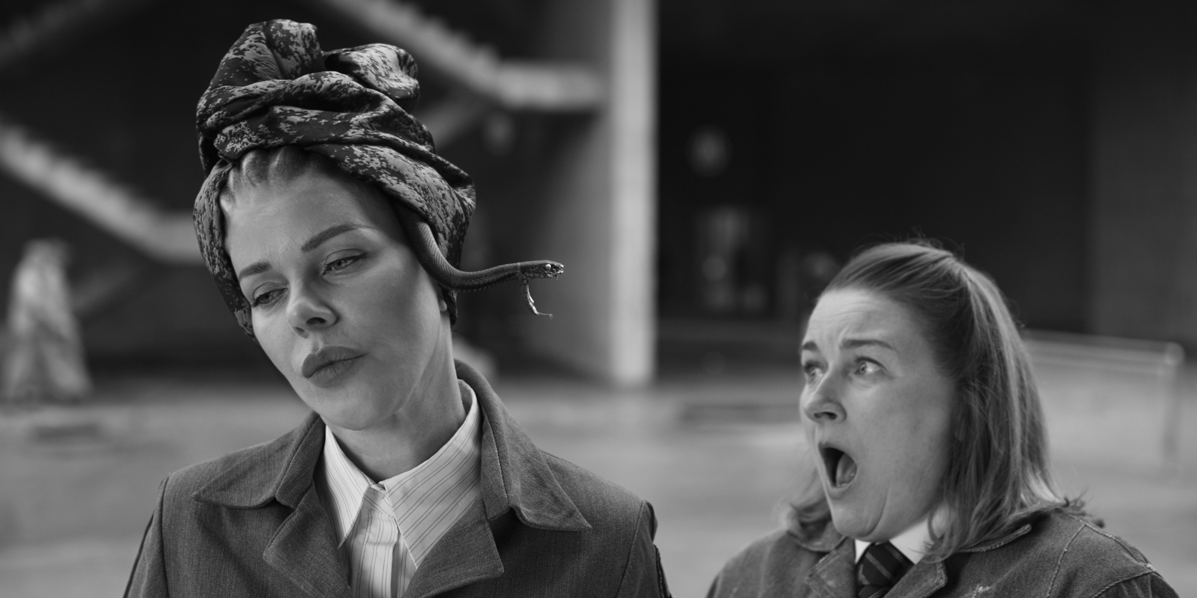 Two women in suits. The woman on the left is looking to her right forlornly, while the woman on the right is looking at the other woman with shock on her face.