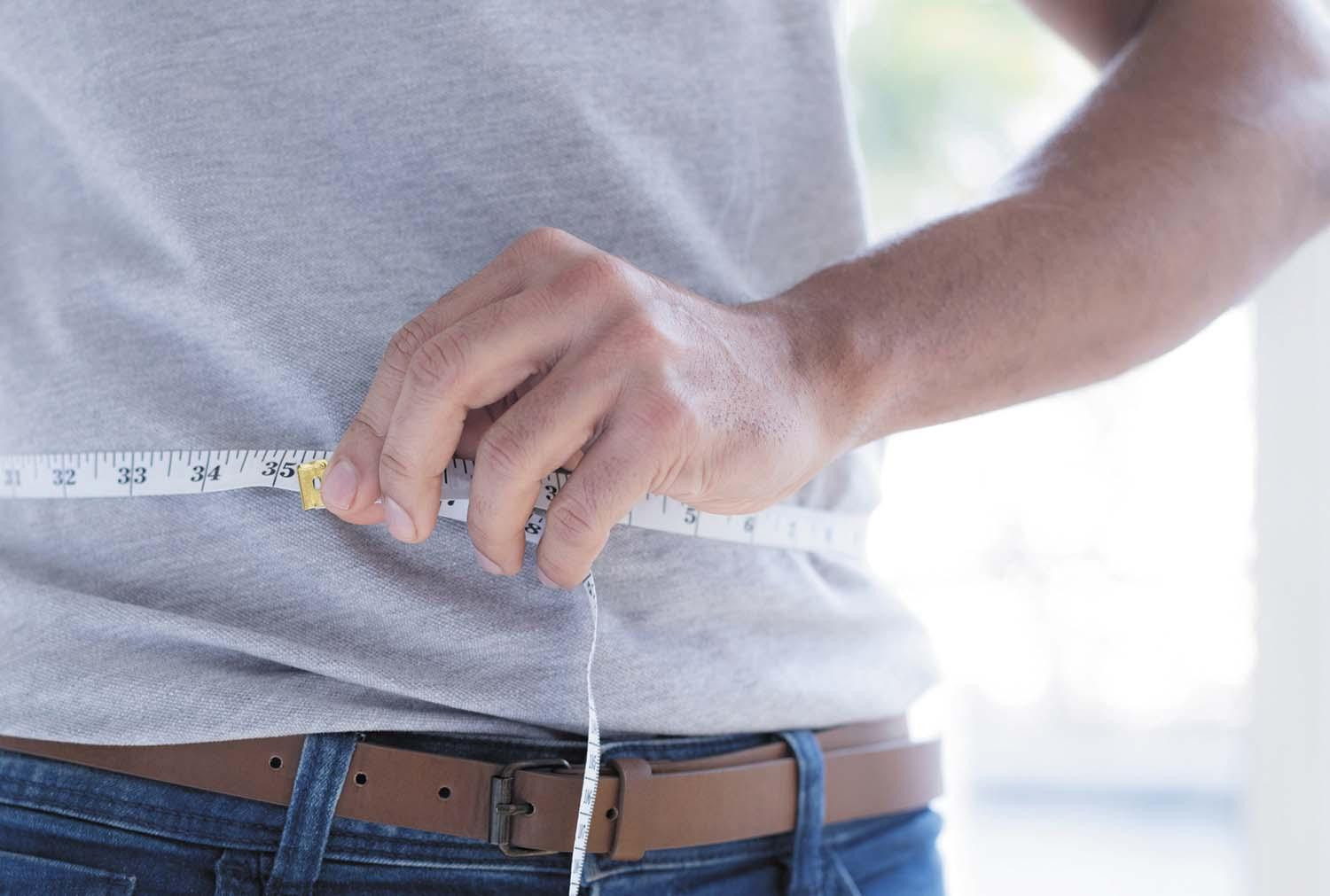 Cropped photo showing the hands of a man measuring his waist with a measuring tape.