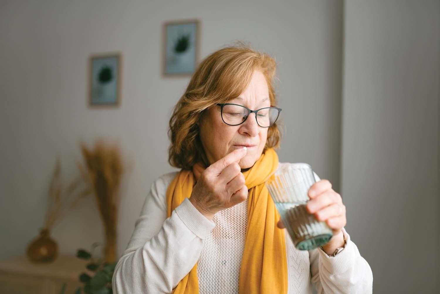 A woman holds a pill near her mouth and holds a glass of water in her other hand, ready to swallow it.