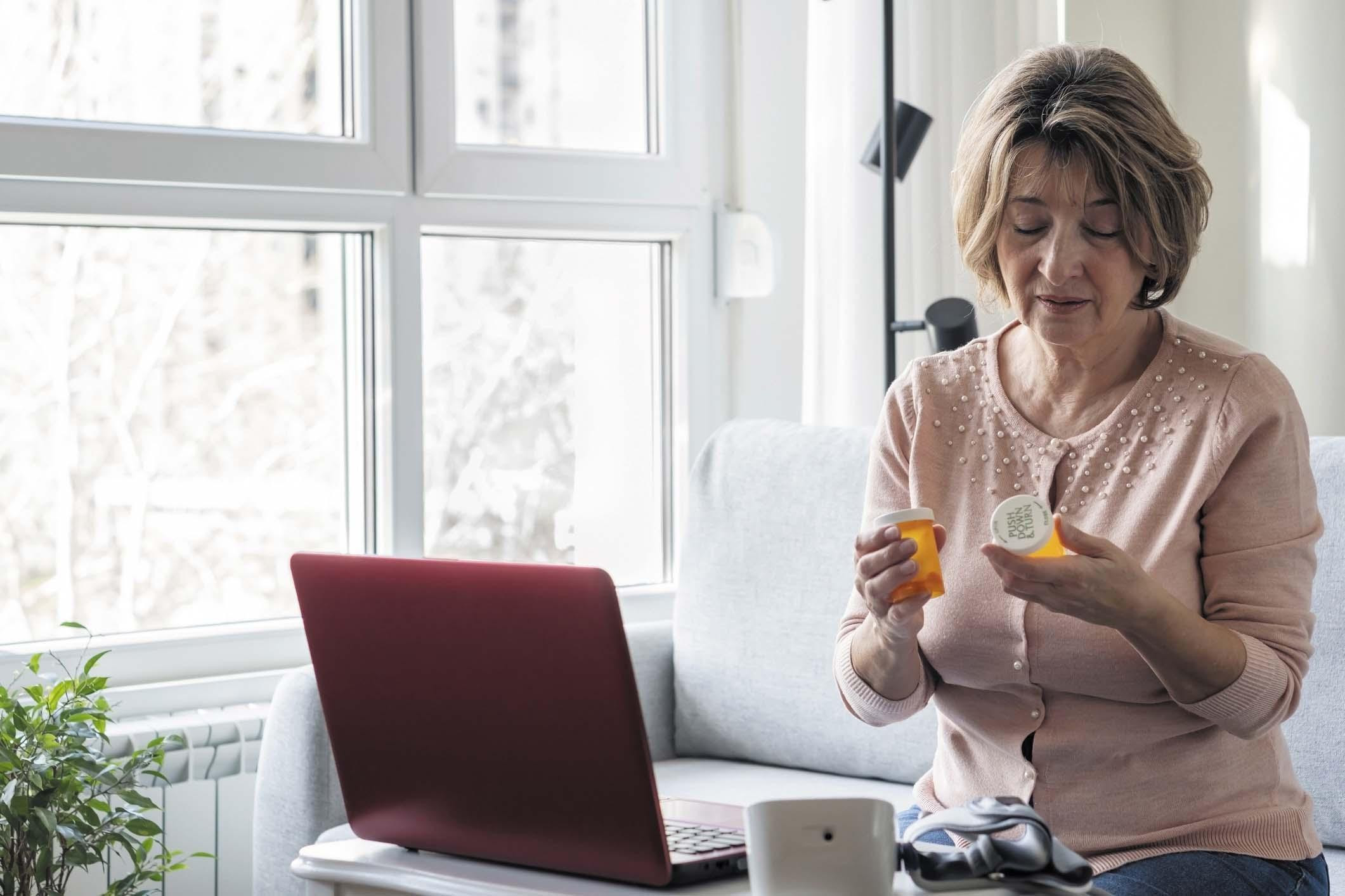 A woman holds a medication bottle and reads the label, standing next to a table with an open laptop on it.
