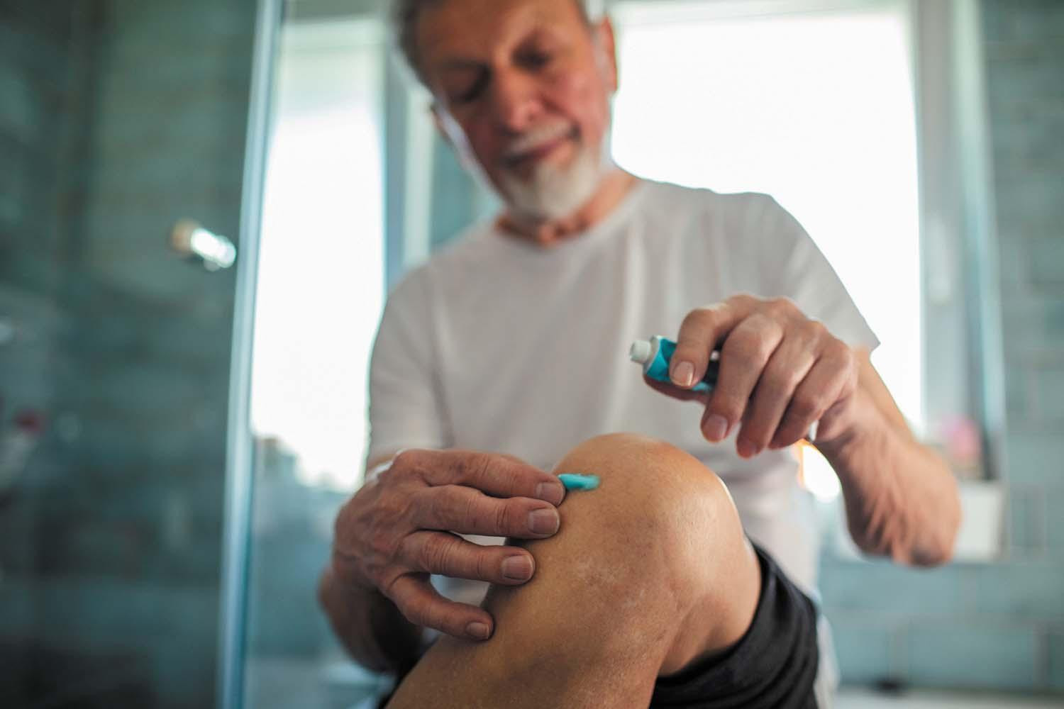 A man applies a topical painkiller to his knee, which is raised in front of him.
