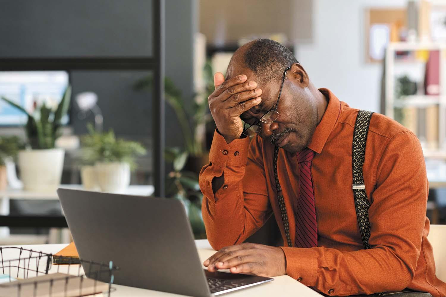 A man sits at a desk with a laptop open in front of him, holding his hand to his forehead.