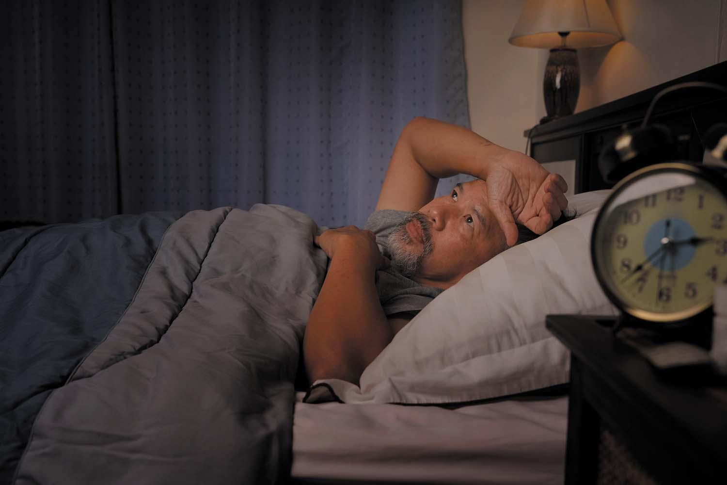 A man awake in bed in a darkened room, holding his wrist on his forehead; in the foreground a clock on the night stand reads 2:38.