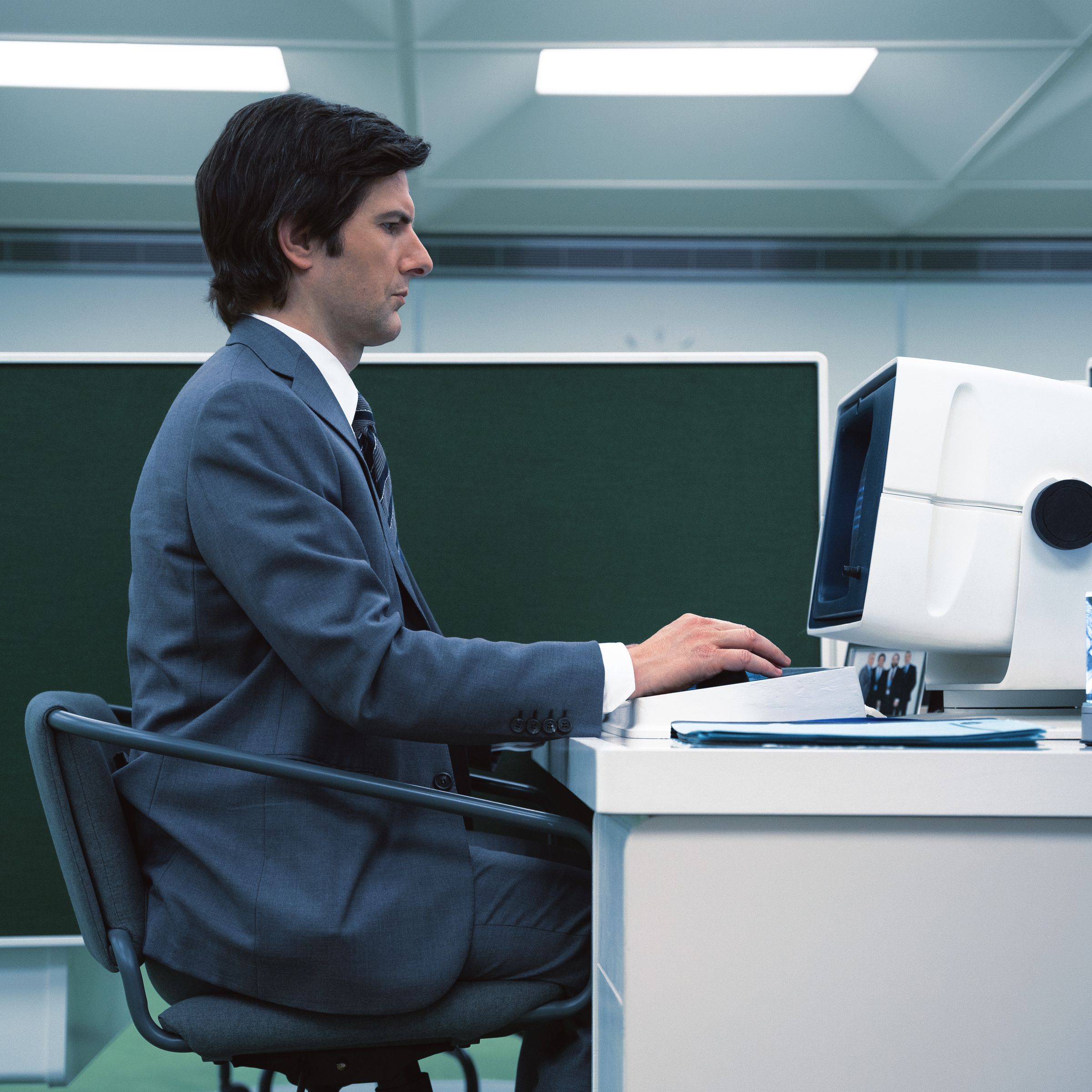 A man in a gray suit sitting at his desk and typing on a computer in a cubicle.